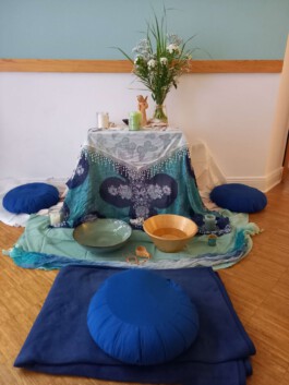 Liebevoll gestalteter Ritualplatz mit brennender Kerze, Blumen, Klangschale und blauen Sitzkissen.