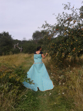 Frau in einem türkisfarbenen Kleid mit gelben Punkten steht auf einer Wiese und berührt die Zweige eines Obstbaums.