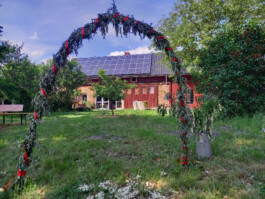 Mit Blumen und Zweigen geschmückter Bogen auf einer Wiese vor einem roten Haus.