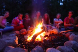 Eine Gruppe sitzt in der Abenddämmerung im Kreis um ein Lagerfeuer, das von großen Natursteinen eingefasst ist.