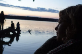 Eine Jugendliche blickt in der Abenddämmerung über einen ruhigen See, während weitere Personen am Ufer sitzen und stehen.