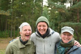 Drei lächelnde Personen stehen eng beieinander auf einer Waldlichtung und haben die Arme umeinandergelegt.