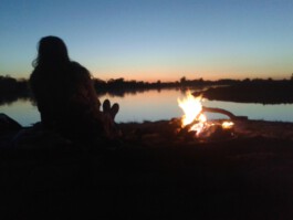 Menschen sitzen bei Sonnenuntergang gemeinsam an einem Feuer am See.