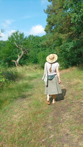 Eine barfüßige Frau mit Strohhut geht auf einem schmalen Weg durch eine sommerliche Waldlandschaft.