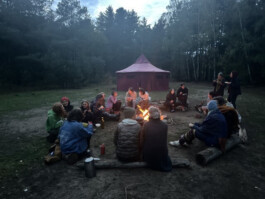 Eine größere Gruppe sitzt in der Dämmerung im Kreis um ein Feuer auf einer Waldlichtung. Im Hintergrund steht ein großes Rundzelt.