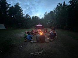Eine Gruppe von Menschen sitzt in der Abenddämmerung gemeinsam um ein Lagerfeuer auf einer Waldlichtung. Im Hintergrund steht ein großes Rundzelt.