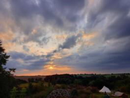 Weite Landschaft mit Wiesen, Bäumen und einem weißen Zelt unter einem wolkenreichen Abendhimmel im Licht der untergehenden Sonne.