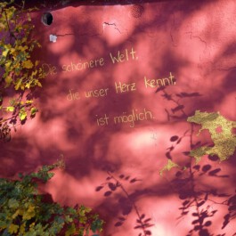 Handschriftlicher Schriftzug „Die schönere Welt, die unser Herz kennt, ist möglich“ auf einer rosafarbenen Wand im Sonnenlicht.