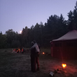Zwei Menschen umarmen sich in der Abenddämmerung auf einer Waldlichtung. Im Hintergrund leuchten ein Feuer und Laternen neben einem großen Zelt.