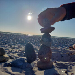 Eine Hand setzt einen weiteren Stein auf einen kleinen Turm aus aufeinanderbalancierten Steinen am Strand. Im Hintergrund scheint die Sonne über dem Meer.